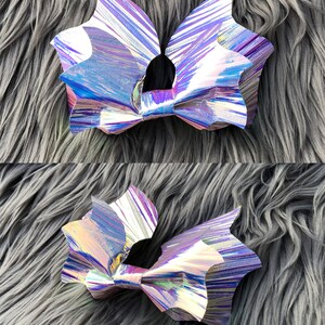 K&ouml;nnte beinhalten: Zwei regenbogenfarbene, holografische Haarb&auml;nder in Fledermausform mit einem Farbschema aus Lila, Blau und Rosa. Die Schleifen liegen auf einer grauen, flauschigen Oberfl&auml;che.