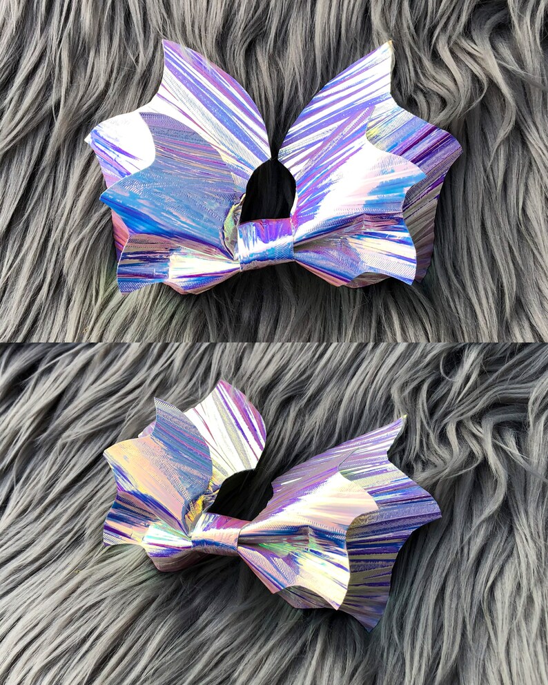 K&ouml;nnte beinhalten: Zwei regenbogenfarbene, holografische Haarb&auml;nder in Fledermausform mit einem Farbschema aus Lila, Blau und Rosa. Die Schleifen liegen auf einer grauen, flauschigen Oberfl&auml;che.