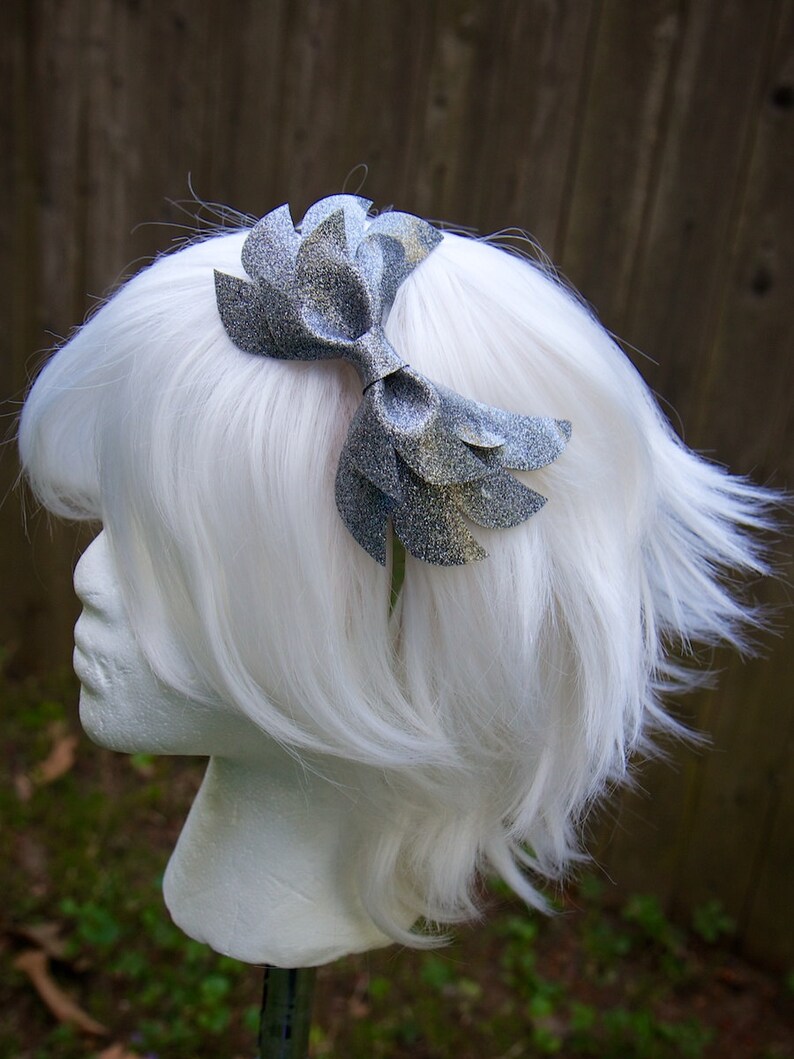 May include: A silver glitter bow headband. The bow is made of multiple layers of fabric and is tied in a knot at the center. The headband is a simple, thin band.