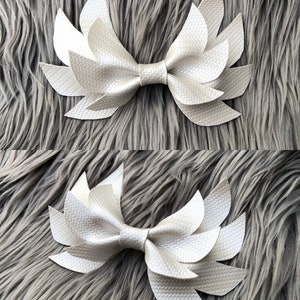 May include: A white faux leather bow with a layered, petal-like design. The bow is made of multiple pieces of leather that are sewn together to create a 3D effect.