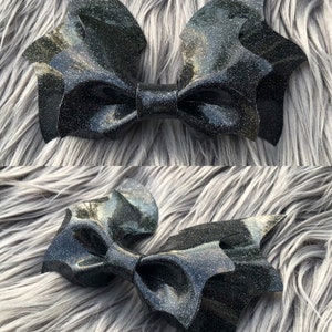 K&ouml;nnte beinhalten: Eine schwarze Glitzer-Fledermausfl&uuml;gel-Schleife. Die Schleife besteht aus einem gl&auml;nzenden schwarzen Stoff mit eingearbeiteten Glitzer. Die Schleife ist in Form von Fledermausfl&uuml;geln gestaltet, mit zwei gro&szlig;en Fl&uuml;geln und einer kleineren mittleren Schleife.