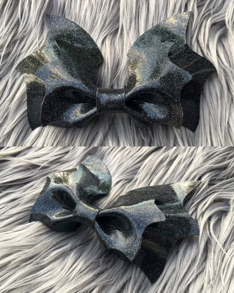 K&ouml;nnte beinhalten: Eine schwarze Glitzer-Fledermausfl&uuml;gel-Schleife. Die Schleife besteht aus einem gl&auml;nzenden schwarzen Stoff mit eingearbeiteten Glitzer. Die Schleife ist in Form von Fledermausfl&uuml;geln gestaltet, mit zwei gro&szlig;en Fl&uuml;geln und einer kleineren mittleren Schleife.