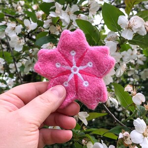 May include: A pink felt cherry blossom flower with white embroidered details. The flower is being held in a hand against a background of a blooming cherry blossom tree.