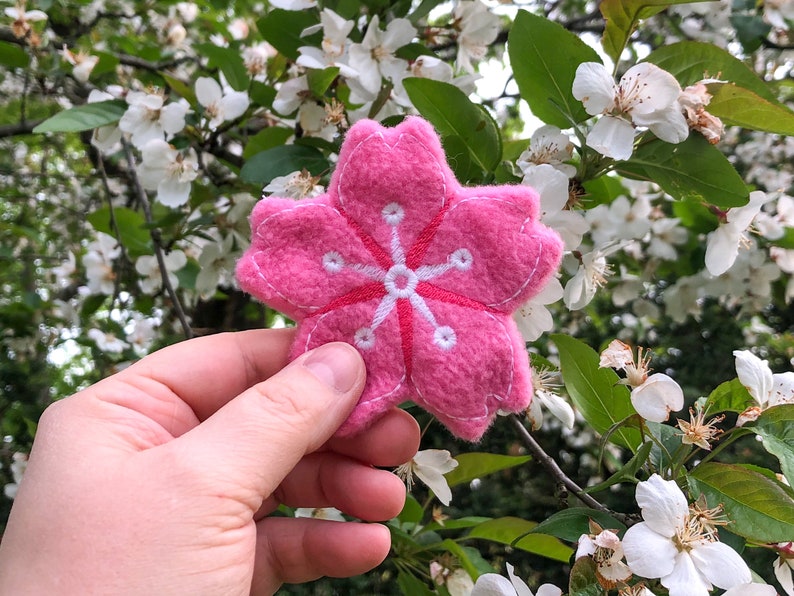 May include: A pink felt cherry blossom flower with white embroidered details. The flower is being held in a hand against a background of a blooming cherry blossom tree.