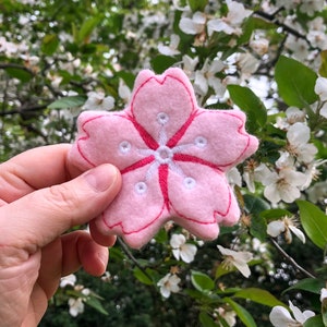 May include: A pink felt cherry blossom flower with white embroidered details. The flower is held in a hand against a background of white cherry blossoms.