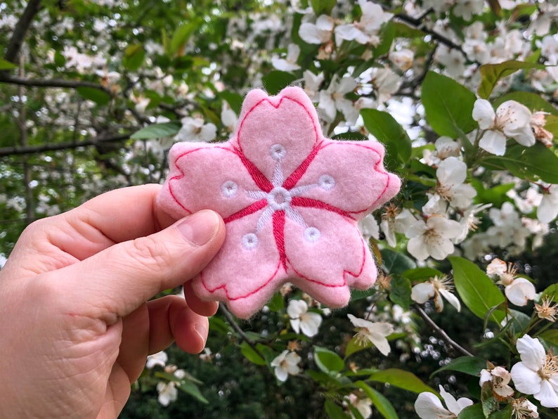 May include: A pink felt cherry blossom flower with white embroidered details. The flower is held in a hand against a background of white cherry blossoms.