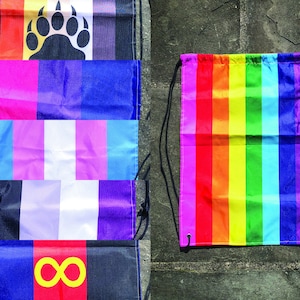 May include: Five different pride flags are displayed on a wall, with a rainbow drawstring bag in the center. The flags include the bear pride flag, the bisexual pride flag, the transgender pride flag, the intersex pride flag, and the infinity pride flag.