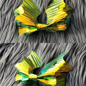 K&ouml;nnte beinhalten: Eine goldene und gr&uuml;ne, irisierende Schleife in Form einer Fledermausfl&uuml;gel. Die Schleife ist aus zerknittertem Folienmaterial und liegt auf einer grauen, flauschigen Oberfl&auml;che.