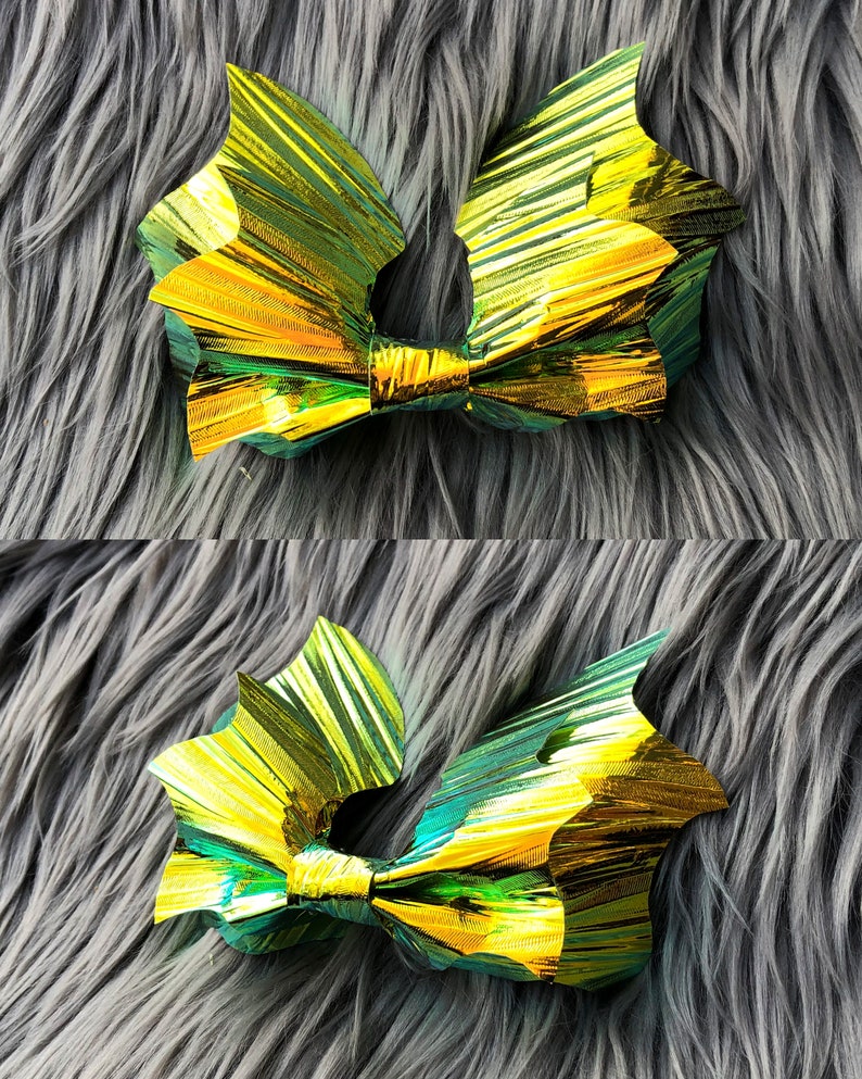 K&ouml;nnte beinhalten: Eine goldene und gr&uuml;ne, irisierende Schleife in Form einer Fledermausfl&uuml;gel. Die Schleife ist aus zerknittertem Folienmaterial und liegt auf einer grauen, flauschigen Oberfl&auml;che.