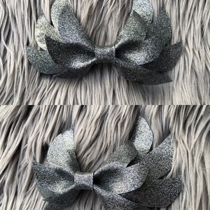 May include: A silver glitter bow with a wing-like design. The bow is made of fabric and has a sparkly finish.