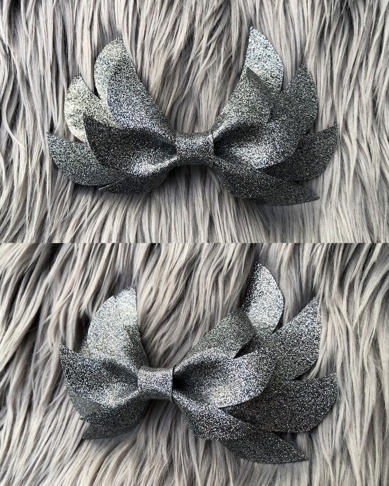 May include: A silver glitter bow with a wing-like design. The bow is made of fabric and has a sparkly finish.