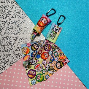 May include: Two Daruma doll-themed keychains with black carabiner clips and a patterned microfiber cloth. The keychains feature colorful designs with the Japanese character for "fortune." The cloth displays Daruma dolls, lucky cats, and floral patterns.