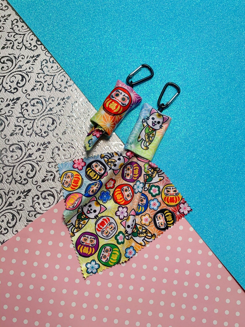 May include: Two Daruma doll-themed keychains with black carabiner clips and a patterned microfiber cloth. The keychains feature colorful designs with the Japanese character for "fortune." The cloth displays Daruma dolls, lucky cats, and floral patterns.