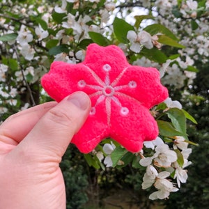 May include: A pink felt flower with a white embroidered design, held in a hand against a background of white blossoms.