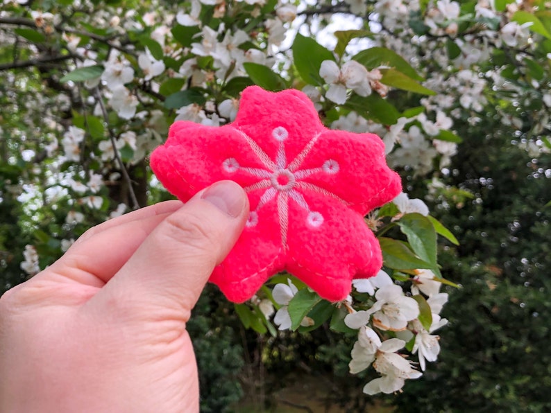 May include: A pink felt flower with a white embroidered design, held in a hand against a background of white blossoms.