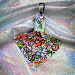 May include: A keychain featuring a Japanese lucky cat design. The keychain has a black carabiner clip and a fabric charm with a waving cat illustration. The charm also displays colorful Daruma dolls and floral patterns.