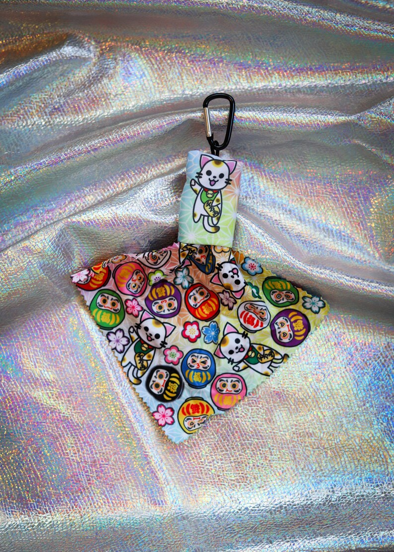 May include: A keychain featuring a Japanese lucky cat design. The keychain has a black carabiner clip and a fabric charm with a waving cat illustration. The charm also displays colorful Daruma dolls and floral patterns.