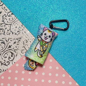 May include: A small, rectangular cat-themed keychain with a black carabiner clip. The keychain features a cartoon lucky cat design with a waving paw, set against a colorful background. A smaller, matching charm is attached.