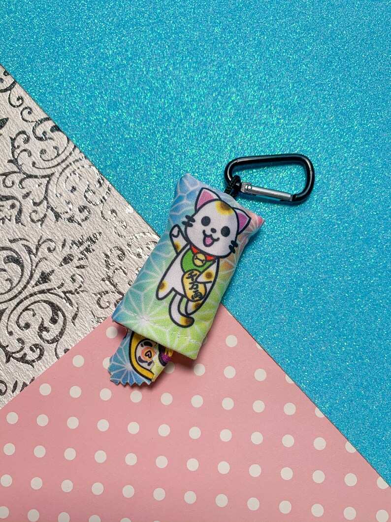 May include: A small, rectangular cat-themed keychain with a black carabiner clip. The keychain features a cartoon lucky cat design with a waving paw, set against a colorful background. A smaller, matching charm is attached.