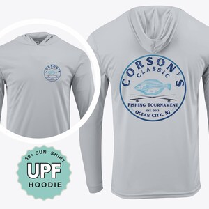 May include: A light gray long-sleeve hooded shirt featuring a blue circular graphic on the back and a smaller matching logo on the front chest. The graphic depicts a fish and two fishing rods with the text Corson's Classic Fishing Tournament Ocean City, NJ Est. 2013. A badge indicates 50+ UPF Sun Shirt Hoodie.