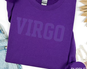 May include: A purple crewneck sweatshirt with the word "VIRGO" printed across the front in a darker shade of purple. The sweatshirt is made of a soft material and has a ribbed collar and cuffs. A small tag is visible on the back of the neck.