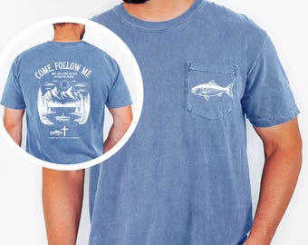 May include: A blue short-sleeve t-shirt with a chest pocket featuring a white fish graphic. The back design displays a mountain lake scene with a boat, a cross, and the text: COME, FOLLOW ME AND I WILL SEND YOU OUT TO FISH FOR PEOPLE MATTHEW 4:19.