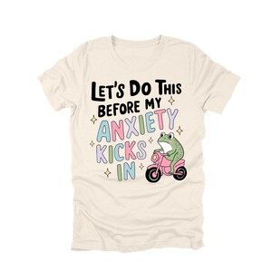 May include: Cream-colored t-shirt with the text "Let's Do This Before My Anxiety Kicks In." A cartoon frog rides a pink scooter. The text and frog are in various colors, including black, blue, green, and pink.