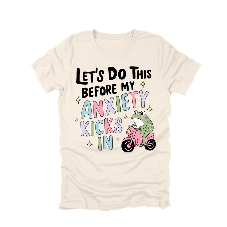 May include: Cream-colored t-shirt with the text "Let's Do This Before My Anxiety Kicks In." A cartoon frog rides a pink scooter. The text and frog are in various colors, including black, blue, green, and pink.