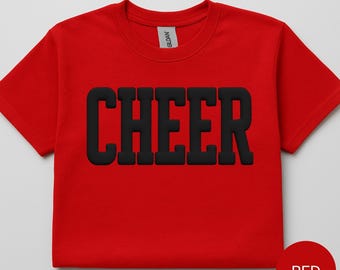 May include: A bright red short-sleeved t-shirt featuring the word CHEER printed in large, bold, black block letters across the center of the chest. The shirt has a crew neckline and a Gildan brand label visible inside the collar area.