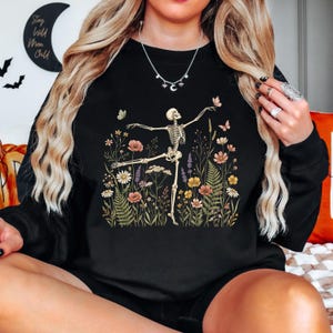 May include: A black crewneck sweatshirt featuring a graphic print of a skeleton dancing amidst a variety of colorful wildflowers, ferns, and small butterflies. The skeleton is positioned in a ballet-like pose, surrounded by detailed botanical illustrations.