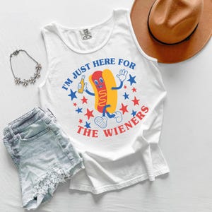 May include: White tank top with a cartoon hot dog graphic holding a beer bottle. The text "I'M JUST HERE FOR THE WIENERS" is printed around the hot dog in blue and red. A brown hat, denim shorts, and a floral necklace are also in the image.