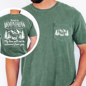 May include: Green t-shirt with a pocket featuring a white mountain and bear graphic. The back of the shirt displays the text "Though the Mountains Move and the Hills Shake, My love will not be removed from you."