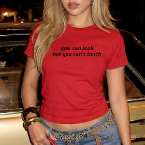 May include: A bright red short-sleeved t-shirt featuring the black text you can look but you can't touch printed across the chest. The garment has a crew neckline and a fitted silhouette, constructed from a solid-colored fabric.