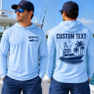 May include: A light blue long-sleeve hooded shirt featuring a dark blue graphic of a fish and the text CUSTOM TEXT on the front, and a larger dark blue graphic of a sport fishing boat and palm trees with the text CUSTOM TEXT on the back.
