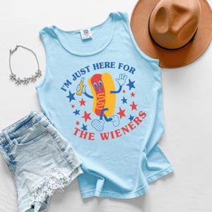 May include: Light blue tank top with the text "I'm just here for the Wieners" and a cartoon hot dog graphic. The shirt is paired with denim shorts, a necklace, and a brown hat.