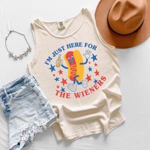 May include: Cream-colored tank top with a cartoon hot dog graphic holding a beer bottle, surrounded by blue and red stars. The text reads "I'M JUST HERE FOR THE WIENERS". The image also includes denim shorts, a necklace, and a brown hat.