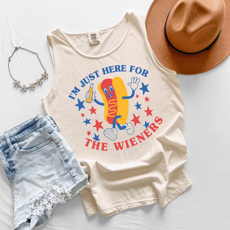 May include: Cream-colored tank top with a cartoon hot dog graphic holding a beer bottle, surrounded by blue and red stars. The text reads "I'M JUST HERE FOR THE WIENERS". The image also includes denim shorts, a necklace, and a brown hat.