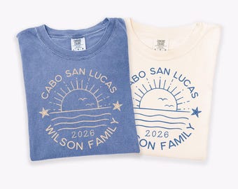 May include: Two crew neck t-shirts, one blue and one cream, each featuring a circular graphic with a sun, waves, stars, and the text Cabo San Lucas, 2026, and Wilson Family. The blue shirt has a light-colored print, while the cream shirt has blue ink.