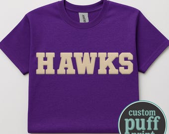 May include: A short-sleeved purple t-shirt featuring the word HAWKS in large, beige, raised puff-print lettering across the chest. The garment has a crew neckline and a Gildan brand label at the inner collar. A circular badge reads custom puff print.