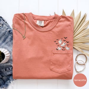 May include: Terracotta-colored t-shirt with a pocket, featuring a design of cartoon ghosts and bats. A gold necklace and bracelets are placed near the shirt. The shirt is a Comfort Colors brand.