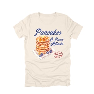 May include: Cream-colored t-shirt featuring a stack of pancakes with butter and syrup, blueberries, and the text "Pancakes & Panic Attacks" in blue. A small red circle says "buy one get one".