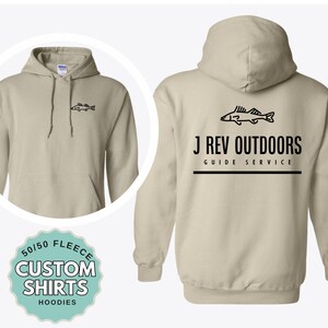 May include: A beige hooded sweatshirt featuring a small black fish graphic on the front left chest. The back displays a larger black fish graphic above the text J REV OUTDOORS GUIDE SERVICE with a horizontal line. A circular badge reads 50/50 FLEECE CUSTOM SHIRTS HOODIES.