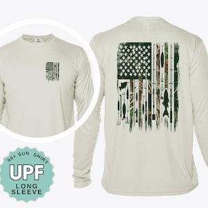 May include: A beige long-sleeve UPF 50+ sun shirt featuring a small American flag graphic on the front left chest and a large American flag design on the back composed of fishing rods, hooks, lures, and fish silhouettes in green and brown tones.
