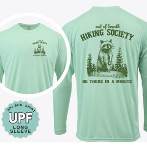 May include: A light green long-sleeve UPF 50+ sun shirt. The front features a small graphic with the text "HIKING SOCIETY" and "BE THERE IN A MINUTE". The back displays a raccoon graphic with the text "out of breath HIKING SOCIETY" and "BE THERE IN A MINUTE".