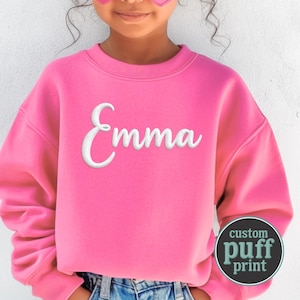 May include: A bright pink long-sleeve sweatshirt featuring the name Emma in white, raised puff-style lettering across the chest. A circular badge in the bottom right corner displays the text custom puff print in white and teal lettering.