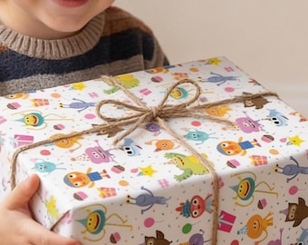May include: A rectangular gift box wrapped in white paper featuring a colorful pattern of small cartoon monsters, stars, and party hats, tied with a simple brown twine bow. The box is held by a smiling child wearing a striped sweater and brown pants.