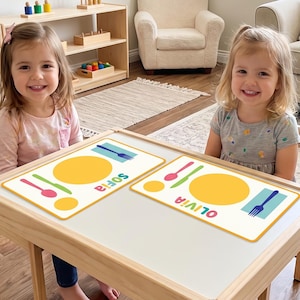 Könnte beinhalten: Zwei rechteckige weiße Platzsets mit gelben kreisförmigen Tellergrafiken, bunten Besteckillustrationen und den Namen Sofia und Olivia in mehrfarbigen Buchstaben, die auf einem Tisch aus hellem Holz platziert sind.