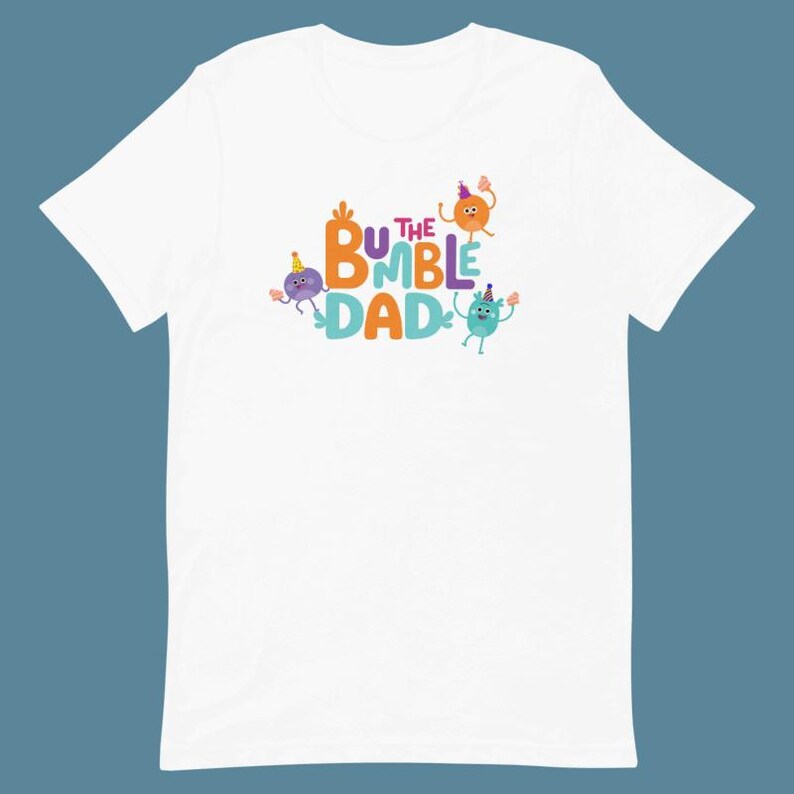 Puede incluir: Camiseta blanca con el texto "BUMBLE DAD" en letras coloridas y personajes de dibujos animados. El dise&ntilde;o presenta figuras naranjas, moradas y turquesas con sombreros de fiesta, a&ntilde;adiendo un toque l&uacute;dico.
