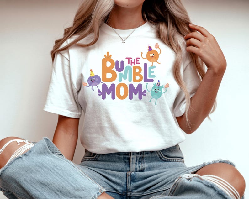 Puede incluir: Camiseta blanca con el texto "The Bumble Mom" en naranja, morado y azul. La camiseta presenta personajes de dibujos animados con gorros de fiesta. La camiseta est&aacute; hecha de un material suave.