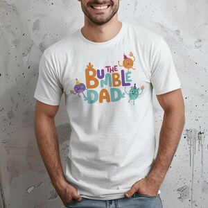 Puede incluir: Camiseta blanca con el texto "The Bumble Dad" en naranja, azul y verde. El dise&ntilde;o incluye personajes de dibujos animados con sombreros de fiesta. La camiseta es de cuello redondo y tiene mangas cortas.
